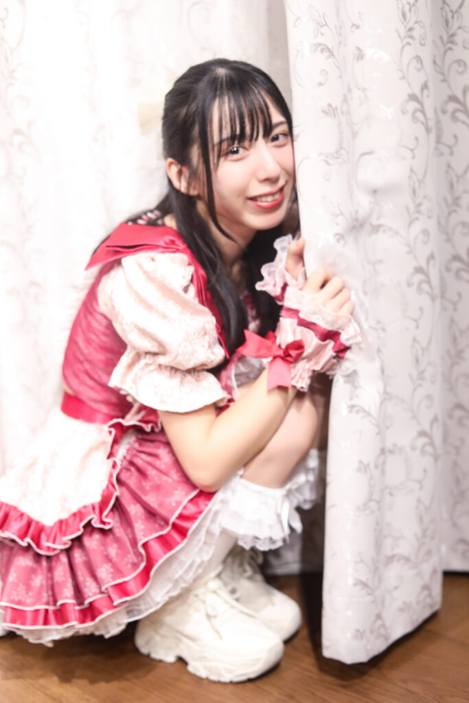 embellir(あんべりー)の水野ひなたちゃんを特典会中に撮影したソロショット。
しゃがんでカーテンの陰に少し隠れるようなポーズで微笑んでいる。とても可愛い。
2025年11月24日 GOTANDA G3 横Fesにて撮影。