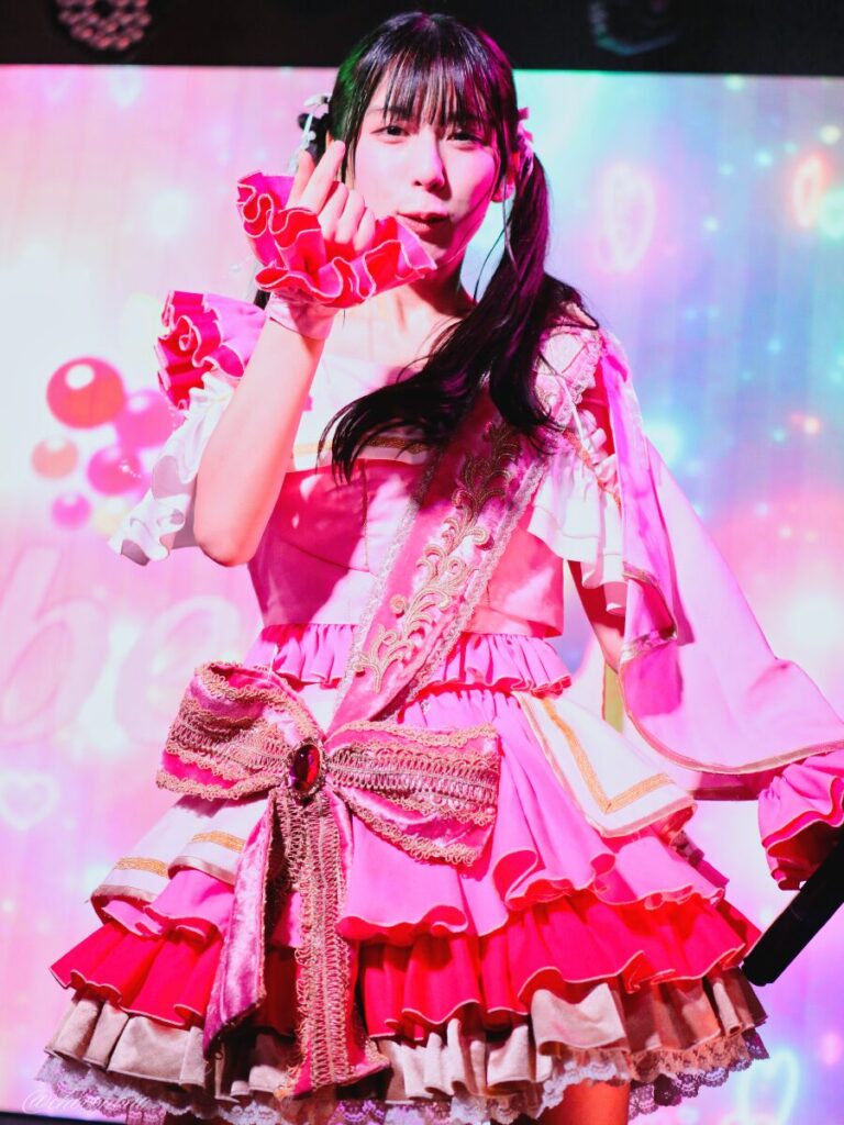 embellir(あんべりー)の水野ひなた(みずのひなた)ちゃんを最前列上手2番辺りからライブ中に撮影した写真。ひなたちゃんはピンクの襷衣装。ツインテールに白いリボンとパールの飾りを着けている。 気が気じゃないのの「球種は直球がいいの」の歌詞の直前、ボールを投げる構えに入っている。 2026年3月3日SHIBUYA ONE5　embellir presents ひなまつりにて撮影。