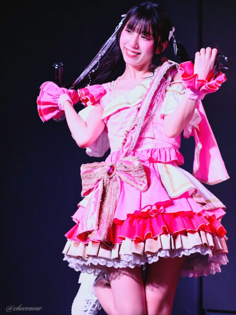 embellir(あんべりー)の水野ひなた(みずのひなた)ちゃんを最前列上手2番辺りからライブ中に撮影した写真。ひなたちゃんはピンクの襷衣装。ツインテールに白いリボンとパールの飾りを着けている。 MC中、髪型と衣装を前にいる人にアピールしている。 2026年3月3日SHIBUYA ONE5　embellir presents ひなまつりにて撮影。