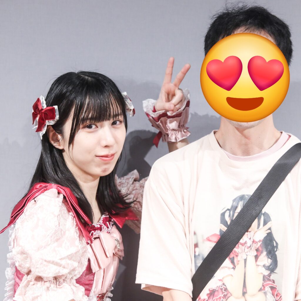 embellir(あんべりー)の水野ひなた(みずのひなた)ちゃんと特典会で撮ってもらったツーショット写真。 ひなたちゃんは毛先を外巻きにして赤いリボンを左右に着けている。 直立した私の髪の横あたりにひなたちゃんがチョキを作って「私が切りました」ポーズ。 2026年3月10日GOTANDA G4　無銭ぷろじぇくと!!にて撮影。