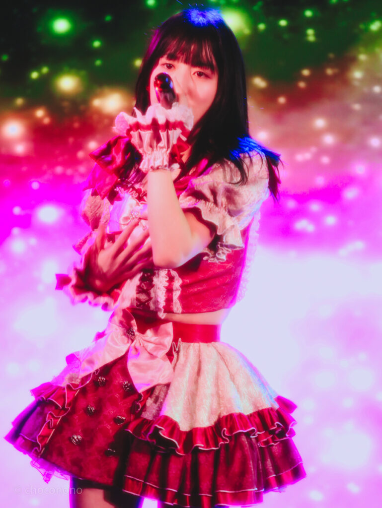 embellir(あんべりー)の水野ひなた(みずのひなた)ちゃんを最前列0番の位置からライブ中に撮影した写真。 ひなたちゃんはストレートヘアーをベースに一束髪を編んでいる。 ベリーベリーの曲中、バックスクリーンに幻想的でカラフルな映像が映っていて、その中で熱唱するひなたちゃんが夢の中にいるよう。 2026年3月15日中野坂上S.U.B Tokyo　妃Fesにて撮影。