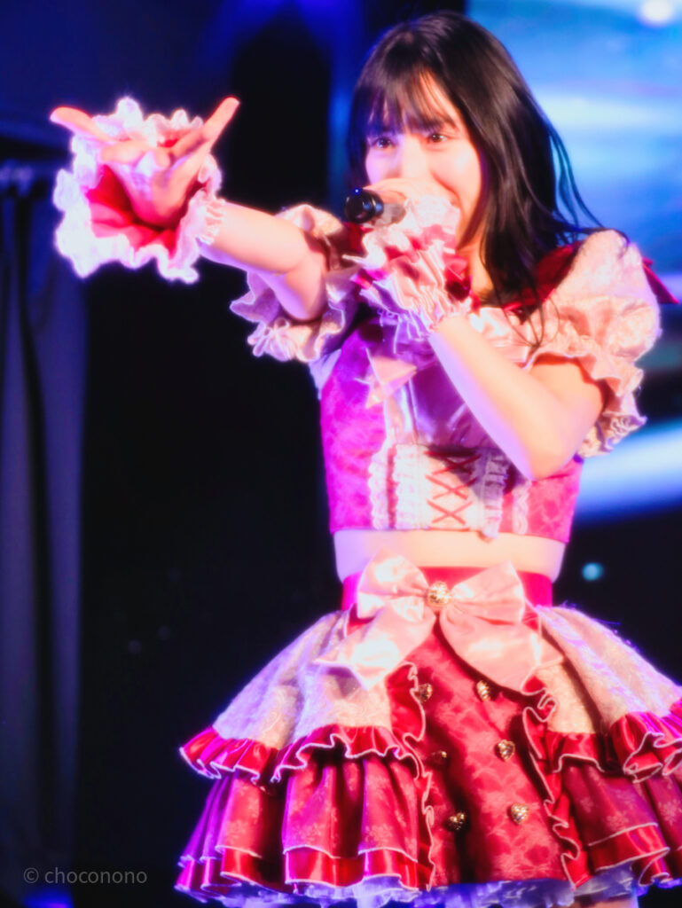 embellir(あんべりー)の水野ひなた(みずのひなた)ちゃんを最前列0番の位置からライブ中に撮影した写真。 ひなたちゃんはストレートヘアーをベースに一束髪を編んでいる。 しっちゃかめっちゃかパラダイスの「みんなといればなんとかなるさ」の歌詞に合わせて手を差しのべるジェスチャーをしている。 2026年3月15日中野坂上S.U.B Tokyo　妃Fesにて撮影。