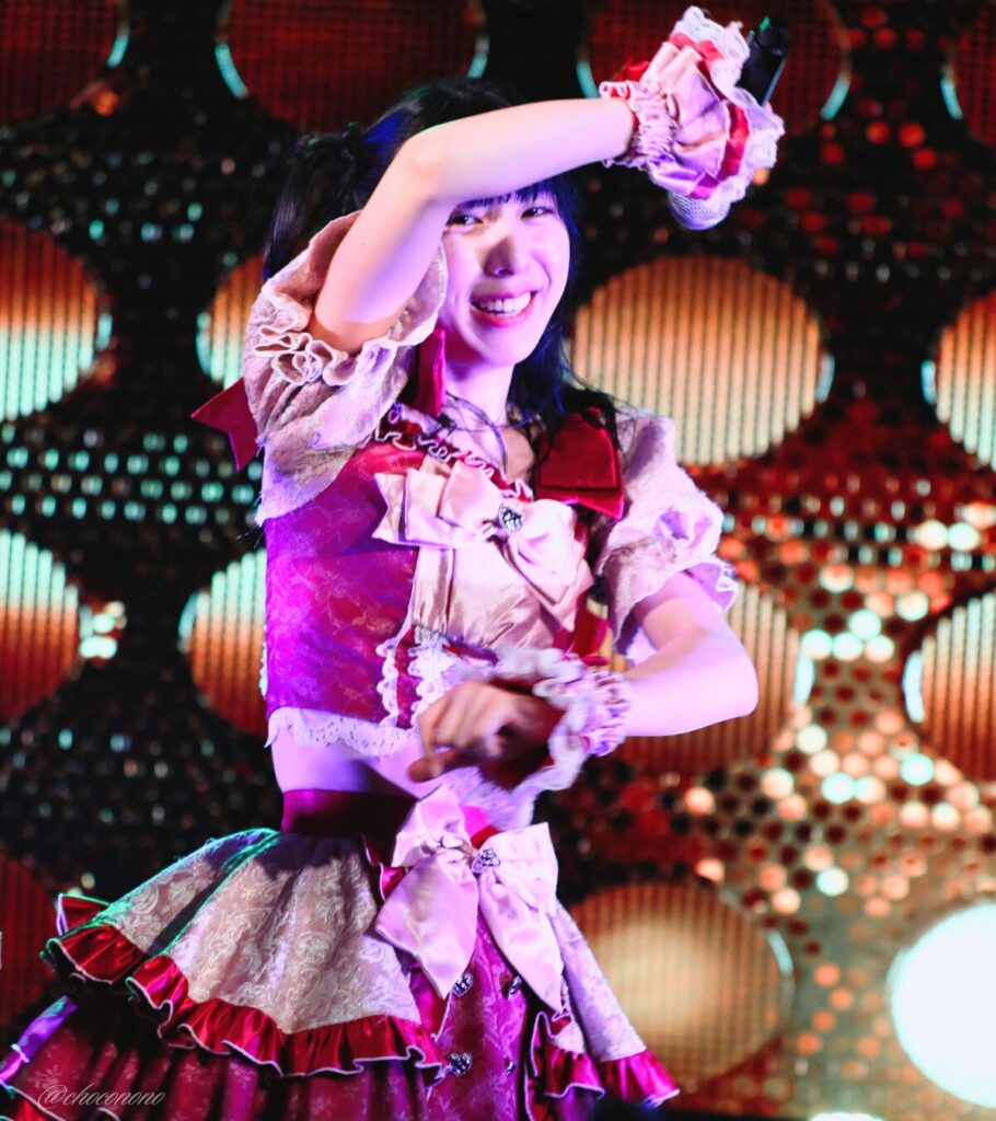 embellir(あんべりー)の水野ひなた(みずのひなた)ちゃんを最前列0番の位置からライブ中に撮影した写真。 ひなたちゃんはハーフツインの根元を輪っかに結んでハムちゃんヘアー。 ダンスの途中、平行に伸ばした右腕と左腕を上下に開いてその間から顔をのぞかせて微笑んでいる。 2026年4月5日中野坂上S.U.B Tokyo 妃Fesにて撮影。