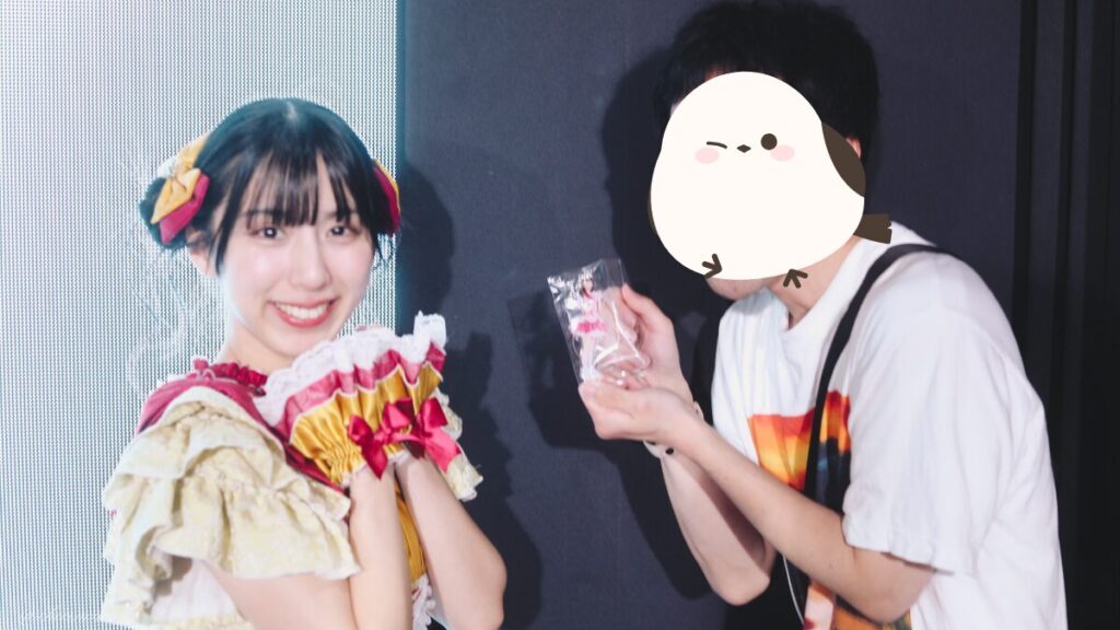 embellir(あんべりー)の水野ひなた(みずのひなた)ちゃんと特典会で撮ってもらったツーショット写真。 ひなたちゃんは普段と衣装を交換してきいろ色の衣装。お団子からちょろちょろとしっぽが出ているような髪型。 私がひなたちゃんのアクキーをアピールし、ひなたちゃんはそのポーズを再現している。 2026年4月9日SHIBUYA ONE5 embellir主催ライブにて撮影。