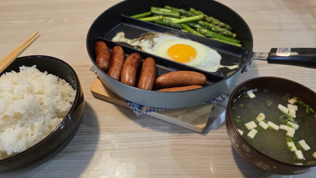 白ご飯、仕切りフライパンを鍋敷きでプレートにした状態で、ウィンナー、目玉焼き、アスパラ炒めが乗っている、お味噌汁