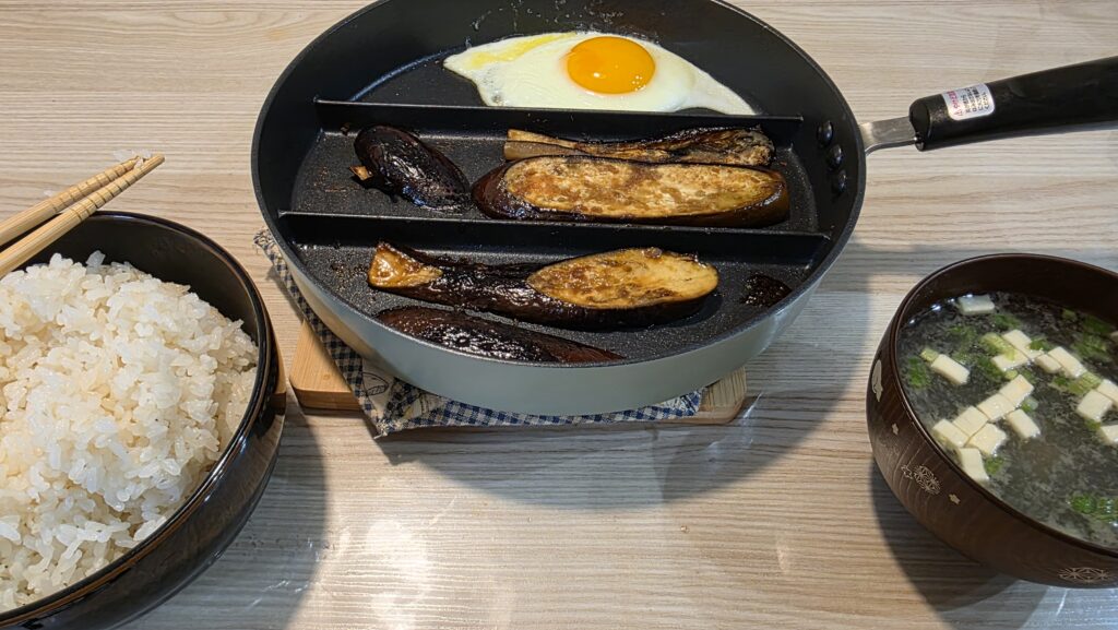 白ご飯、仕切りフライパンに乗った目玉焼きとなすの生姜焼き、お味噌汁