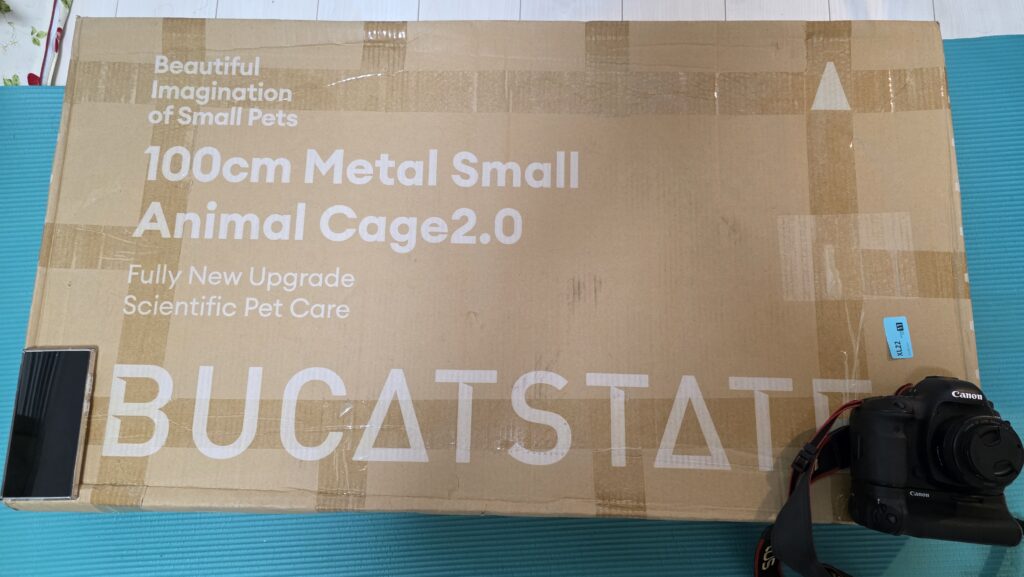 BUCATSTATEの100cmケージが届いた状態。大きさ比較のためにスマートフォンと一眼レフカメラを乗せてみている