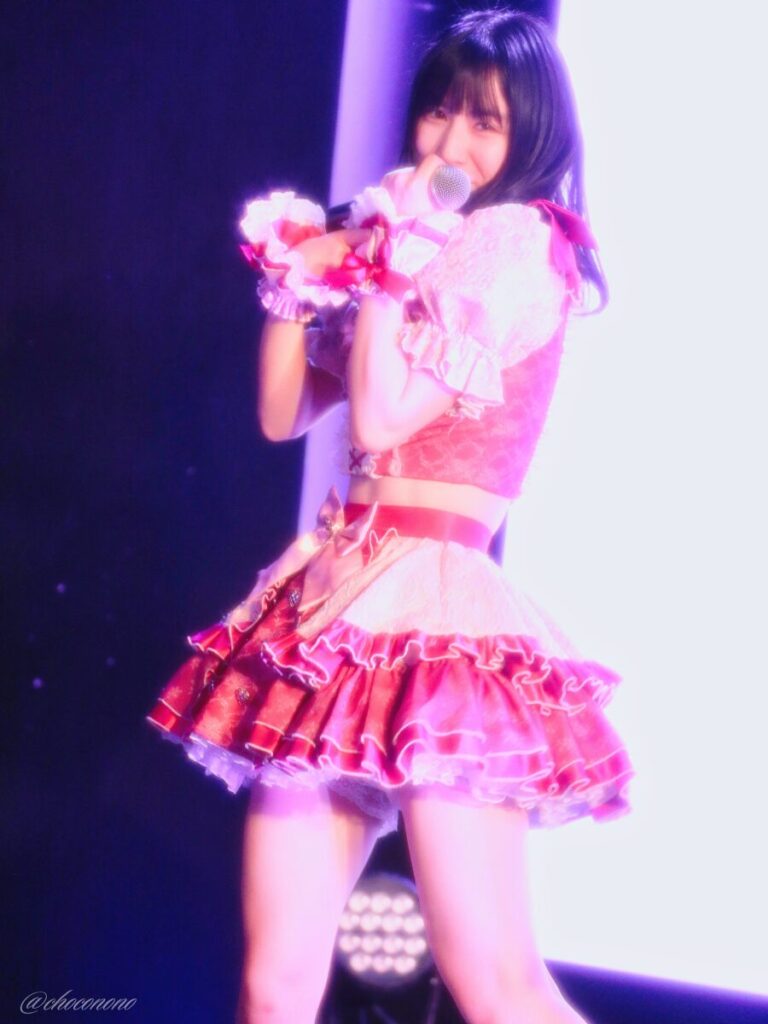 embellir(あんべりー)の水野ひなた(みずのひなた)ちゃんを最前列0番の位置からライブ中に撮影した写真。 ひなたちゃんはストレートヘアーをベースに一束髪を編んでいる。 マイクで少し顔を隠すようにしながらいたずらっぽく微笑んでいる。 2026年3月15日中野坂上S.U.B Tokyo　妃Fesにて撮影。