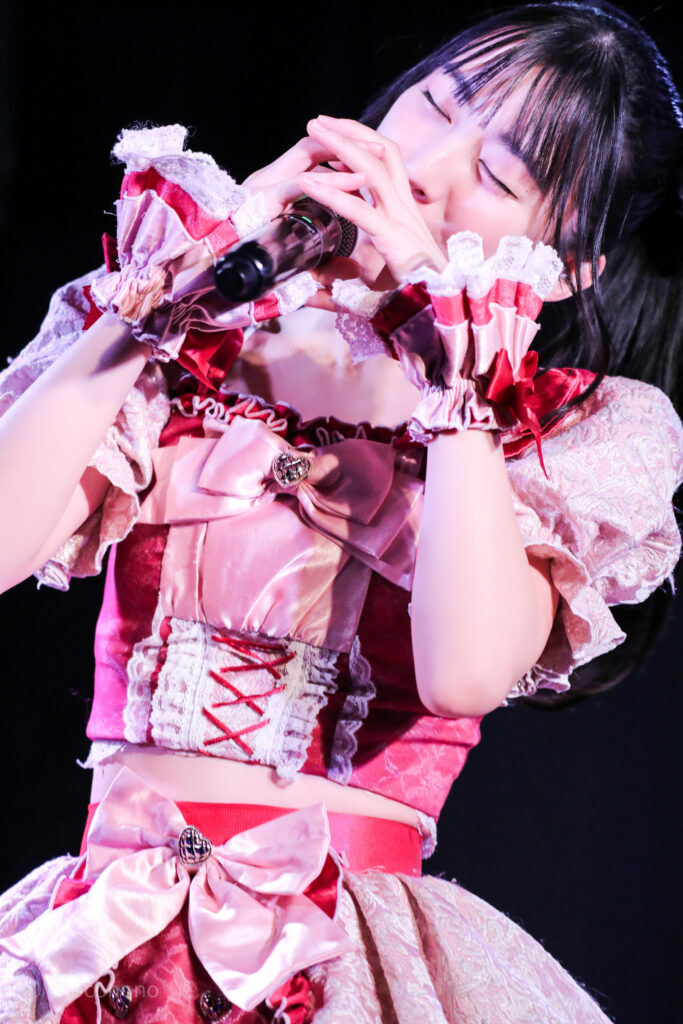 embellir(あんべりー)の水野ひなた(みずのひなた)ちゃんを最前0番からライブ中に撮影した写真。 お気に入りの紺色のリボンにポニーテール。MCでのメンバー甘夏あもちゃんの「あもと甘いおしるこ食べよう」に合わせておしるこを飲むようなジェスチャーをしている。 2026年1月18日中野坂上S.U.B Tokyo IDOL ADVENTUREにて撮影