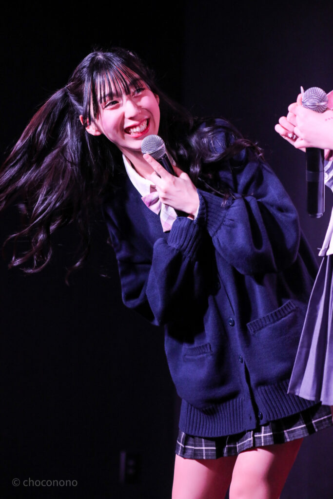 embellir(あんべりー)の水野ひなた(みずのひなた)ちゃんをライブ中に撮影した写真。ピンクリボンの制服に紺のセーター。 メンバーと顔を見合わせてにこにこと笑っている。 2026年2月13日SHIBUYA ONE5　embellir presents VALENTINE LIVEにて撮影。