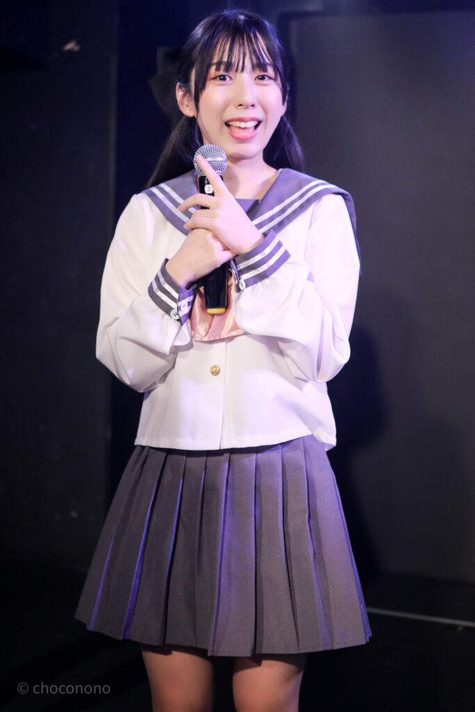 embellir(あんべりー)の水野ひなた(みずのひなた)ちゃんを最前列0番の位置からライブ中に撮影した写真。ピンクリボンのセーラー服にツインテールと大きな黒リボン。 カメラに向かってにこにこと微笑んでくれている。 2026年2月14日新宿DHNoA　IDOL FABRIC Valentineにて撮影。