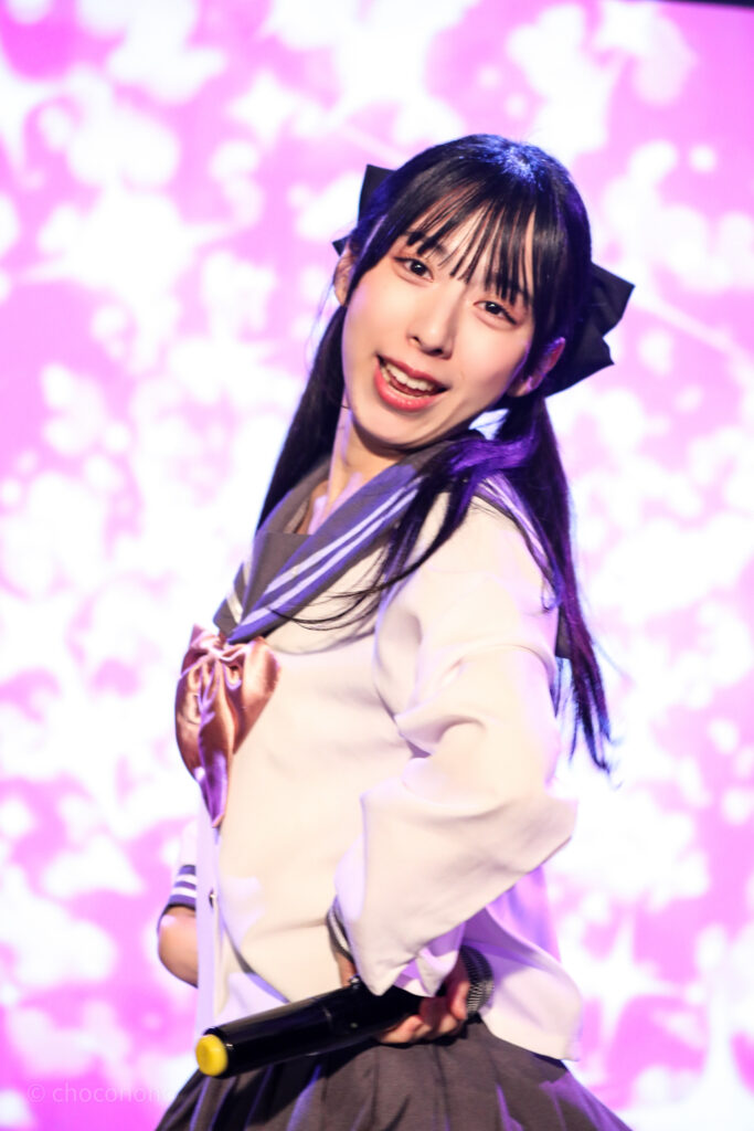 embellir(あんべりー)の水野ひなた(みずのひなた)ちゃんを最前列0番の位置からライブ中に撮影した写真。ピンクリボンのセーラー服にツインテールと大きな黒リボン。 ダンスの最中、カメラに向かってにこにこと微笑んでくれている。 2026年2月14日新宿DHNoA　IDOL FABRIC Valentineにて撮影。