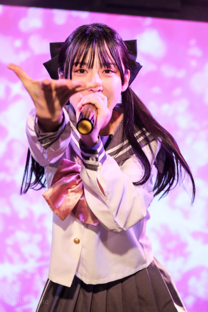 embellir(あんべりー)の水野ひなた(みずのひなた)ちゃんを最前列0番の位置からライブ中に撮影した写真。ピンクリボンのセーラー服にツインテールと大きな黒リボン。 こちらに向かって手を差し伸べてくれている。 2026年2月14日新宿DHNoA　IDOL FABRIC Valentineにて撮影。