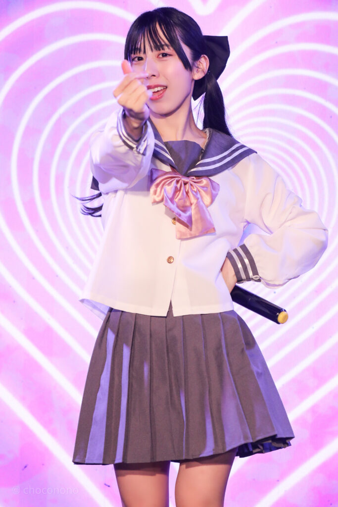 embellir(あんべりー)の水野ひなた(みずのひなた)ちゃんを最前列0番の位置からライブ中に撮影した写真。ピンクリボンのセーラー服にツインテールと大きな黒リボン。 ハートLupinの「私のハートLupin」の歌詞に合わせて指ハートを作っている。 2026年2月14日新宿DHNoA　IDOL FABRIC Valentineにて撮影。