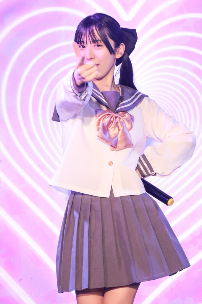 embellir(あんべりー)の水野ひなた(みずのひなた)ちゃんを最前列0番の位置からライブ中に撮影した写真。ピンクリボンのセーラー服にツインテールと大きな黒リボン。 ハートLupinの「私のハートLupin」の歌詞に合わせて指で銃を作ってこちらを撃ち抜いている。 2026年2月14日新宿DHNoA　IDOL FABRIC Valentineにて撮影。