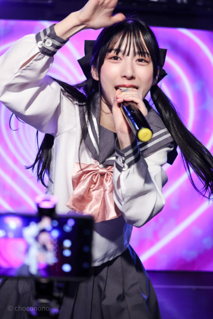 embellir(あんべりー)の水野ひなた(みずのひなた)ちゃんを最前列0番の位置からライブ中に撮影した写真。ピンクリボンのセーラー服にツインテールと大きな黒リボン。 ハートLupinの「じっと見張っててね」の歌詞に合わせて人を探しているジェスチャー。 2026年2月14日新宿DHNoA　IDOL FABRIC Valentineにて撮影。