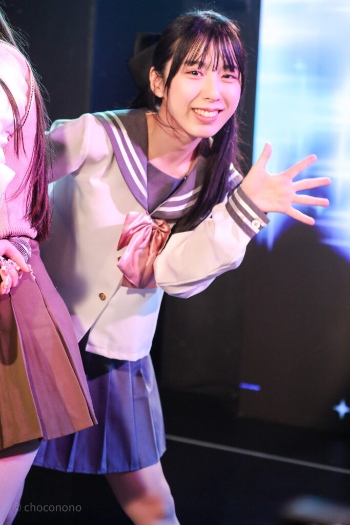 embellir(あんべりー)の水野ひなた(みずのひなた)ちゃんを最前列0番の位置からライブ中に撮影した写真。ピンクリボンのセーラー服にツインテールと大きな黒リボン。 前にいるメンバーの陰からひょっこり出てきたようなポーズ。 2026年2月14日新宿DHNoA　IDOL FABRIC Valentineにて撮影。