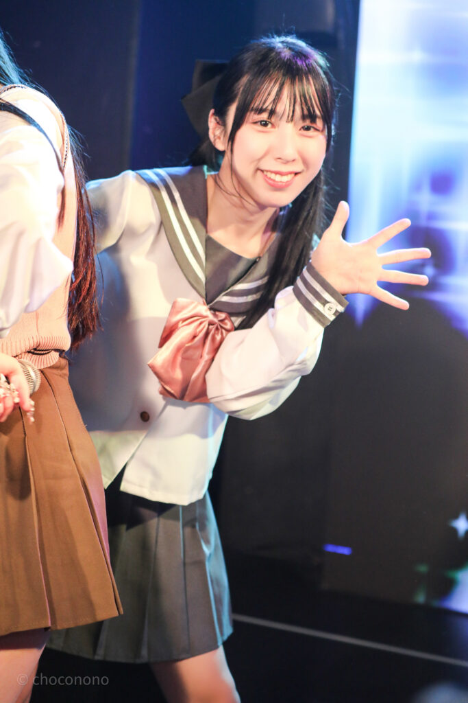 embellir(あんべりー)の水野ひなた(みずのひなた)ちゃんを最前列0番の位置からライブ中に撮影した写真。ピンクリボンのセーラー服にツインテールと大きな黒リボン。 前にいるメンバーの陰からひょっこり出てきたようなポーズ。 2026年2月14日新宿DHNoA　IDOL FABRIC Valentineにて撮影。