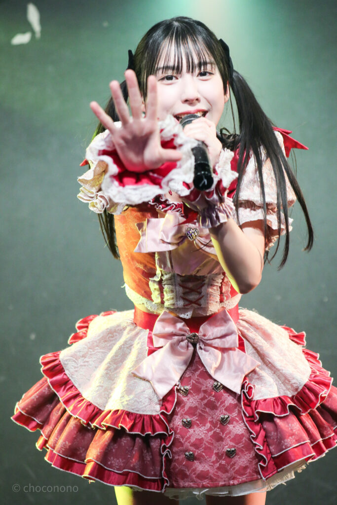 embellir(あんべりー)の水野ひなた(みずのひなた)ちゃんを最前列0番の位置からライブ中に撮影した写真。 黒いリボンでツインテールにしている。 カメラに向かって手のひらを差し出しながら微笑んでいる。 2026年2月21日GOTANDA G6　無銭ぷろじぇくと!!にて撮影。