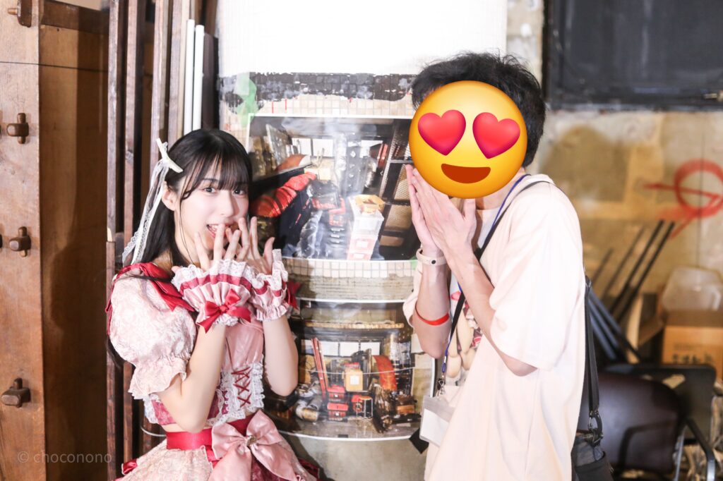 embellir(あんべりー)の水野ひなた(みずのひなた)ちゃんと特典会で撮ってもらったツーショット写真。 ひなたちゃんは巻いた髪に白いリボンを着けている。 倉庫でアイドルと二人きりというシチュエーションでドキッとしている表情。 2026年2月23日東京キネマ倶楽部　IDOL大博覧会にて撮影。