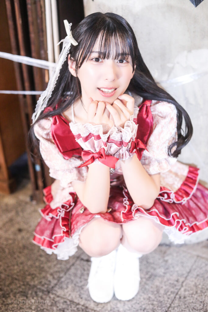 embellir(あんべりー)の水野ひなた(みずのひなた)ちゃんを特典会で撮らせてもらったソロショット写真。 ひなたちゃんは巻いた髪に白いリボンを着けている。 しゃがんでカメラ目線で微笑んでくれている。 2026年2月23日東京キネマ倶楽部　IDOL大博覧会にて撮影。