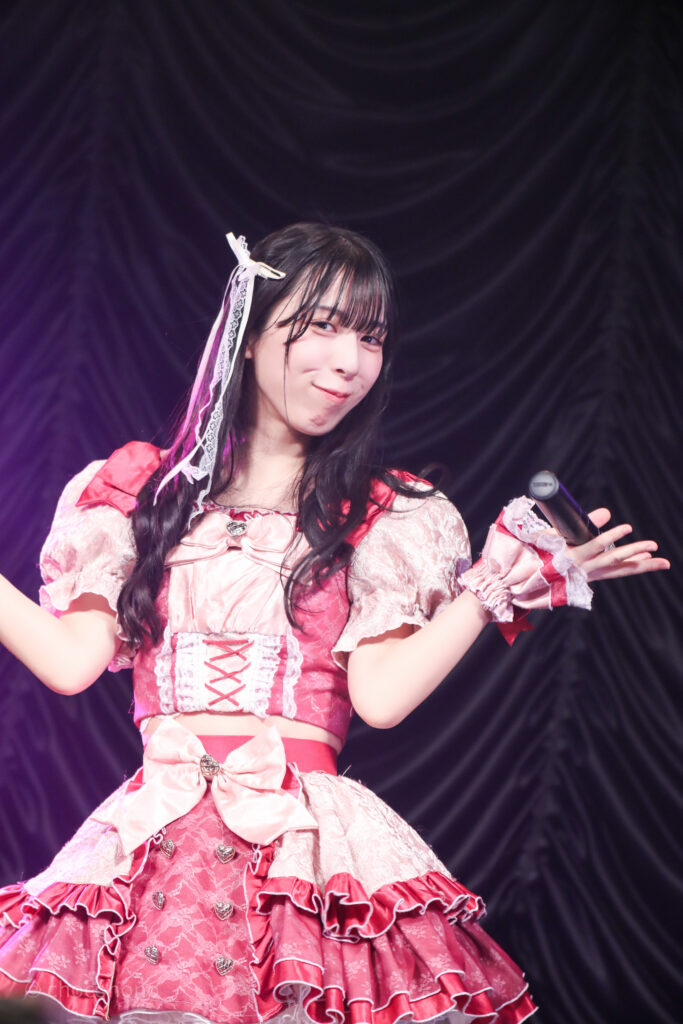 embellir(あんべりー)の水野ひなた(みずのひなた)ちゃんを最前列0番の位置からライブ中に撮影した写真。 ひなたちゃんは巻いた髪に白いリボンを着けている。 得意げな表情でこちらを見ながら微笑んでいる。 2026年2月23日東京キネマ倶楽部　IDOL大博覧会にて撮影。