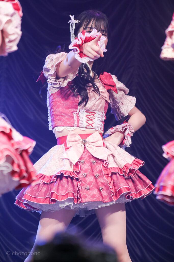 embellir(あんべりー)の水野ひなた(みずのひなた)ちゃんを最前列0番の位置からライブ中に撮影した写真。 ひなたちゃんは巻いた髪に白いリボンを着けている。 ときめきフルスロットルの「君を見つけたの」の歌詞の部分でこちらを指さしてくれている。その手で隠れて表情は見えないが、見えている目元の感じから、多分にこにこしている。 2026年2月23日東京キネマ倶楽部　IDOL大博覧会にて撮影。