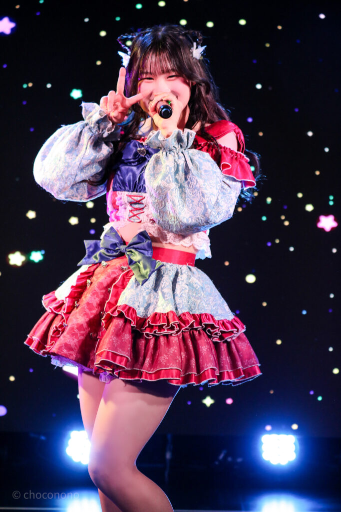 embellir(あんべりー)の恋羽あゆう(こはねあゆう)ちゃんを最前列0番の位置から撮影したライブ写真。 あゆうちゃんは髪で猫耳を作っている。 にこにこと微笑みながらこちらにピースをしつつ、マイクを構えて歌唱している。 2026年2月22日中野坂上S.U.B Tokyo　妃Fesにて撮影。