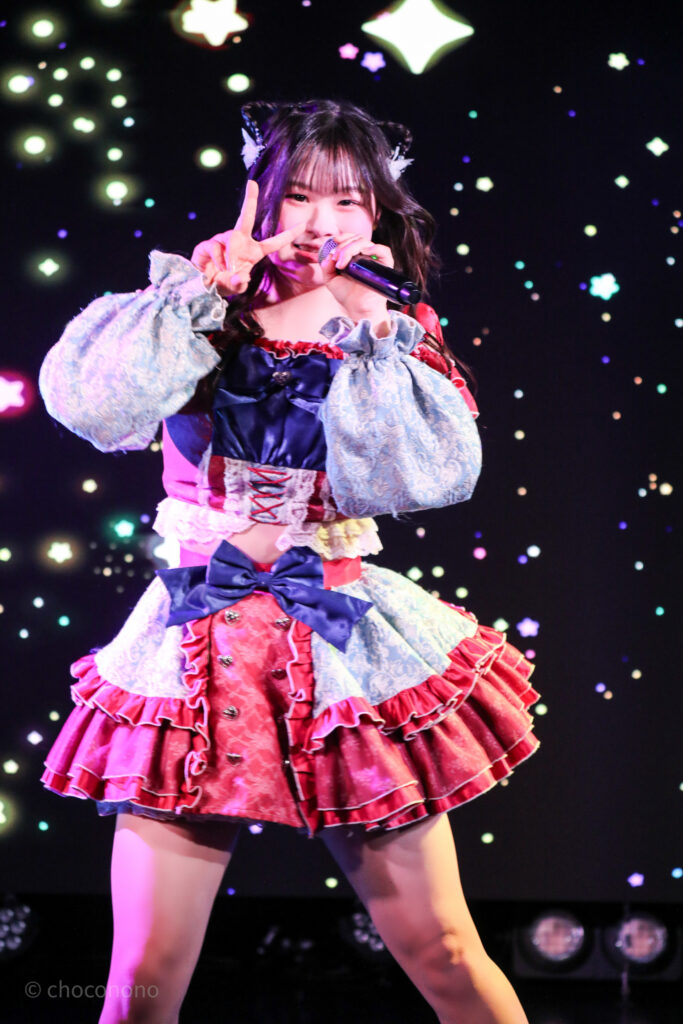 embellir(あんべりー)の恋羽あゆう(こはねあゆう)ちゃんを最前列0番の位置から撮影したライブ写真。 あゆうちゃんは髪で猫耳を作っている。 にこにこと微笑みながらこちらにピースをしつつ、マイクを構えて歌唱している。 2026年2月22日中野坂上S.U.B Tokyo　妃Fesにて撮影。