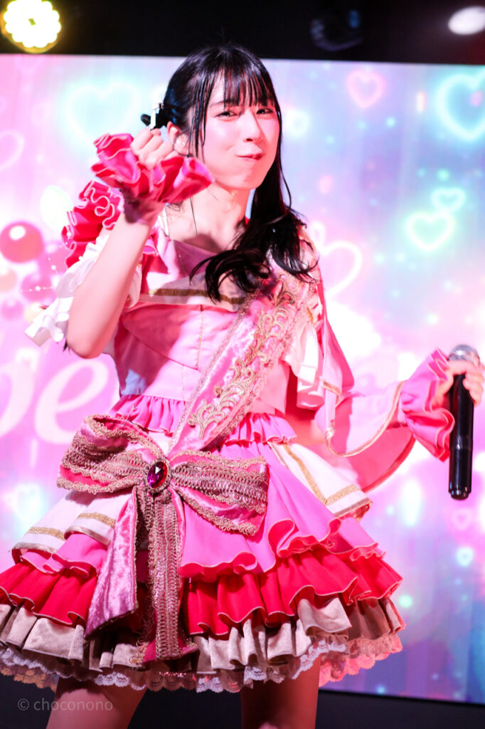 embellir(あんべりー)の水野ひなた(みずのひなた)ちゃんを最前列上手2番辺りからライブ中に撮影した写真。ひなたちゃんはピンクの襷衣装。ツインテールに白いリボンとパールの飾りを着けている。 得意げな表情でこちらを見つめながら手に持った何かをﾁｬｶﾁｬｶ振るジェスチャーをしている。 2026年3月3日SHIBUYA ONE5　embellir presents ひなまつりにて撮影。