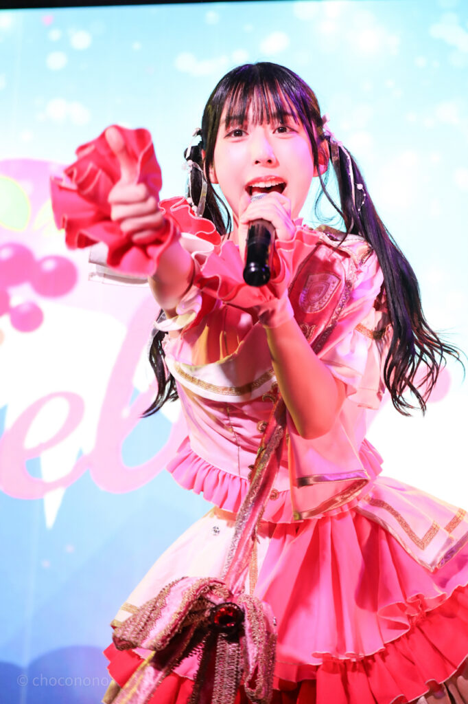 embellir(あんべりー)の水野ひなた(みずのひなた)ちゃんを最前列上手2番辺りからライブ中に撮影した写真。 ひなたちゃんはピンクの襷衣装。ツインテールに白いリボンとパールの飾りを着けている。 前方に向けてサムズアップしながら歌唱している。 2026年3月3日SHIBUYA ONE5　embellir presents ひなまつりにて撮影。