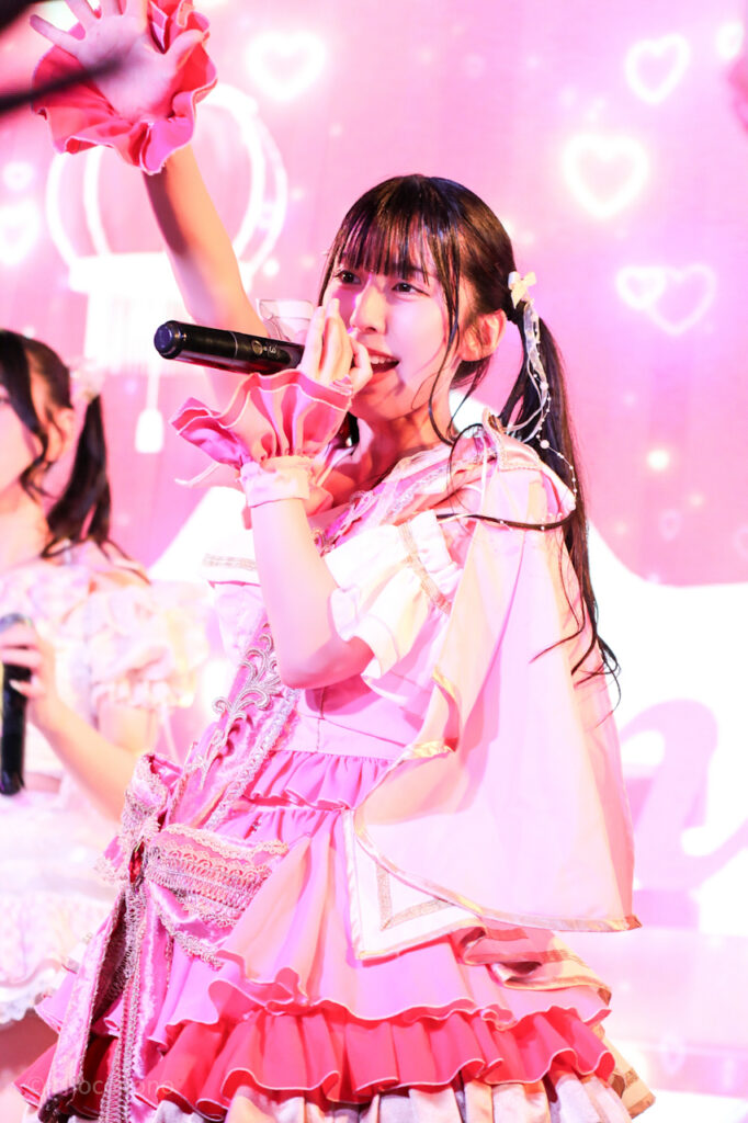 embellir(あんべりー)の水野ひなた(みずのひなた)ちゃんを最前列上手2番辺りからライブ中に撮影した写真。ひなたちゃんはピンクの襷衣装。ツインテールに白いリボンとパールの飾りを着けている。 気が気じゃないのの「ハレルヤ待ってるや」の歌詞に合わせて手のひらを頭上に挙げている。隣の人が同じように掲げた手が、遠近法でハイタッチのようになっている。 2026年3月3日SHIBUYA ONE5　embellir presents ひなまつりにて撮影。