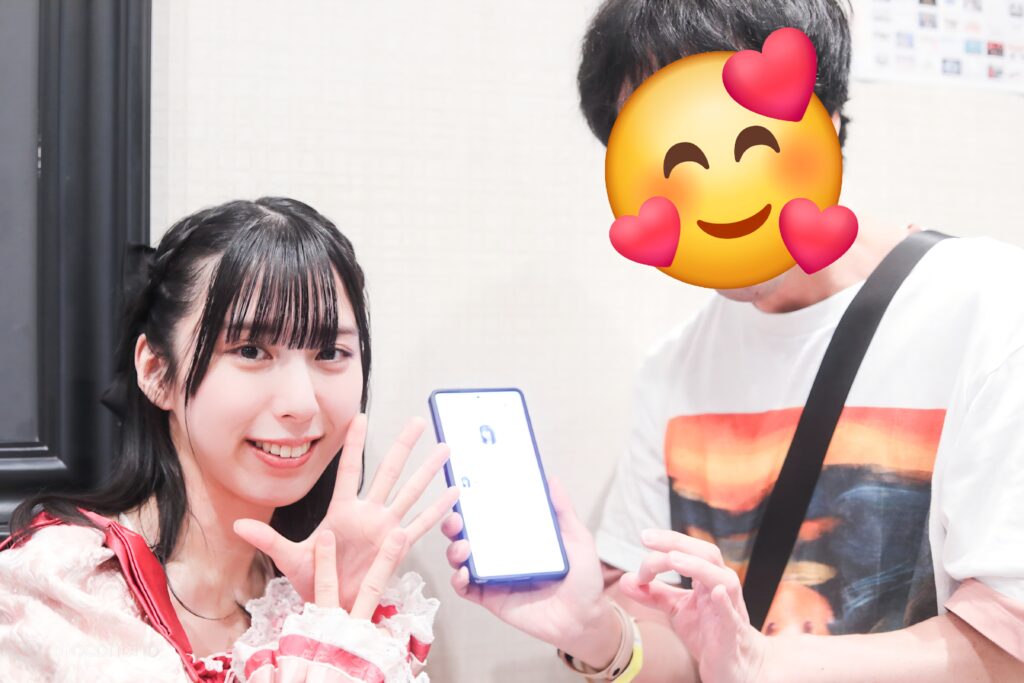 embellir(あんべりー)の水野ひなた(みずのひなた)ちゃんと特典会で撮ってもらったツーショット写真。 ひなたちゃんは編み込みに黒いリボンを着けている。 出会って700日記念日だったので記念日管理アプリと、二人で7と0を手で作っている。 2026年3月7日GOTANDA G6　無銭ぷろじぇくと!!にて撮影。