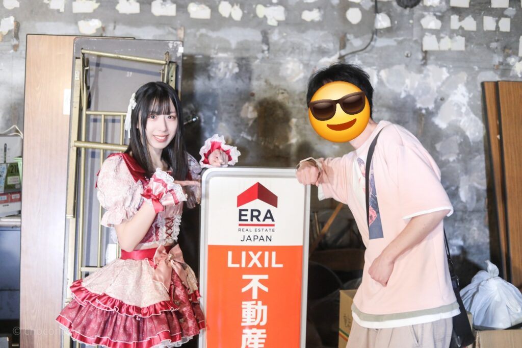 embellir(あんべりー)の水野ひなた(みずのひなた)ちゃんと特典会で撮ってもらったツーショット写真。 ひなたちゃんはストレートヘアーに白いリボンを着けている。 LIXIL不動産と書かれた看板を二人でアピールしている。 2026年3月8日新世紀ホール　iCOLONY iDOL LiVEにて撮影。