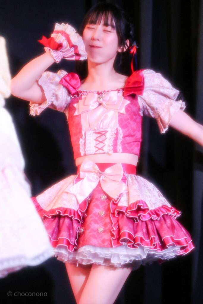 embellir(あんべりー)の水野ひなた(みずのひなた)ちゃんを最前列下手2番あたりから撮ったライブ写真。 真ん中あたりでツインテールにして赤と青のリボンで結んでいる。 プレイフルのラスト、サムズアップした右手を顔の前に置いてにっこり笑っている。 2026年3月14日SHIBUYA ONE5　遥乃ももか×恋羽あゆうおたんじょうびかいにて撮影