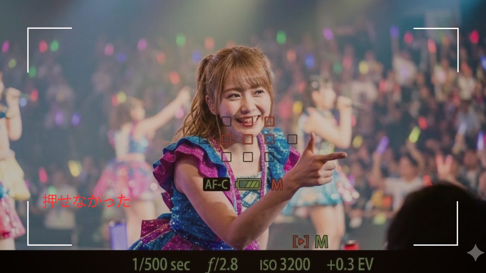 ファインダーから覗いた先にはアイドルがライブパフォーマンスをしている。目の前でピントも合ってシャッターチャンスになるはずが、アイドルの視線も指さしも誰か違う人の方を向いている。 「押せなかった」というテキストが埋め込まれている。