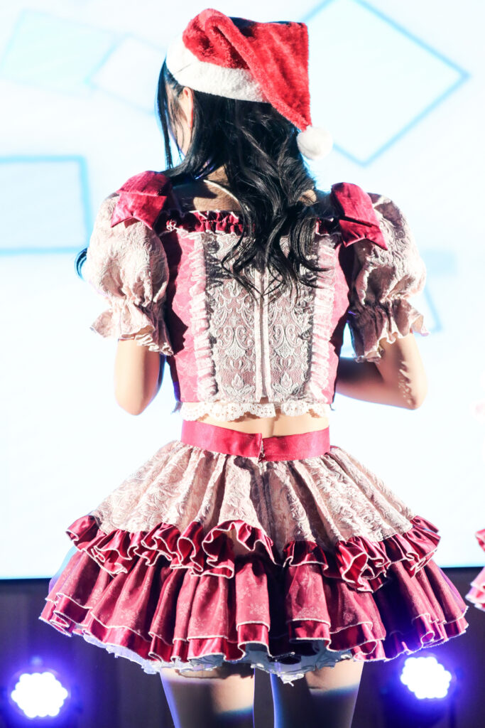 embellir(あんべりー)の水野ひなたちゃんをライブ中に撮影した後ろ姿の写真。
この日はクリスマスイベントということでひなたちゃんはサンタの帽子をかぶっている。
帽子の下から見える後ろ髪は巻かれていてとても大人っぽい魅力を醸し出している。
2025年12月14日　としま区民センター多目的ホール　IDOL SODA Stream!!にて撮影。