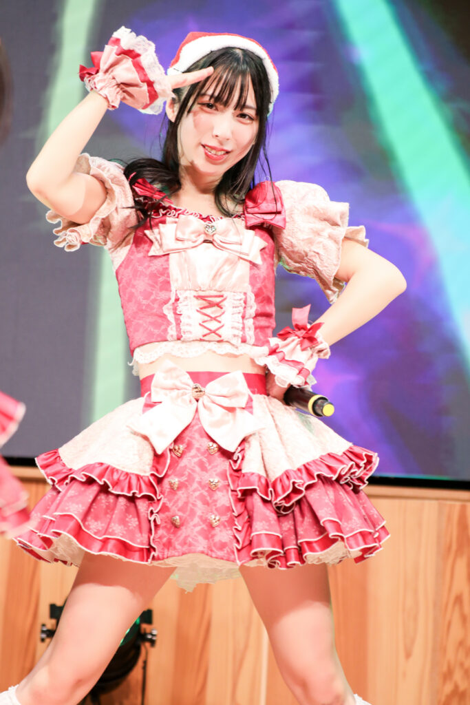 embellir(あんべりー)の水野ひなたちゃんをライブ中に撮影した写真。
この日はクリスマスイベントということでひなたちゃんはサンタの帽子をかぶっている。
こちらを真っ直ぐ見つめながら顔の前で手をひさしのようにして「見つけた」のポーズをしてくれている。
2025年12月14日　としま区民センター多目的ホール　IDOL SODA Stream!!にて撮影。