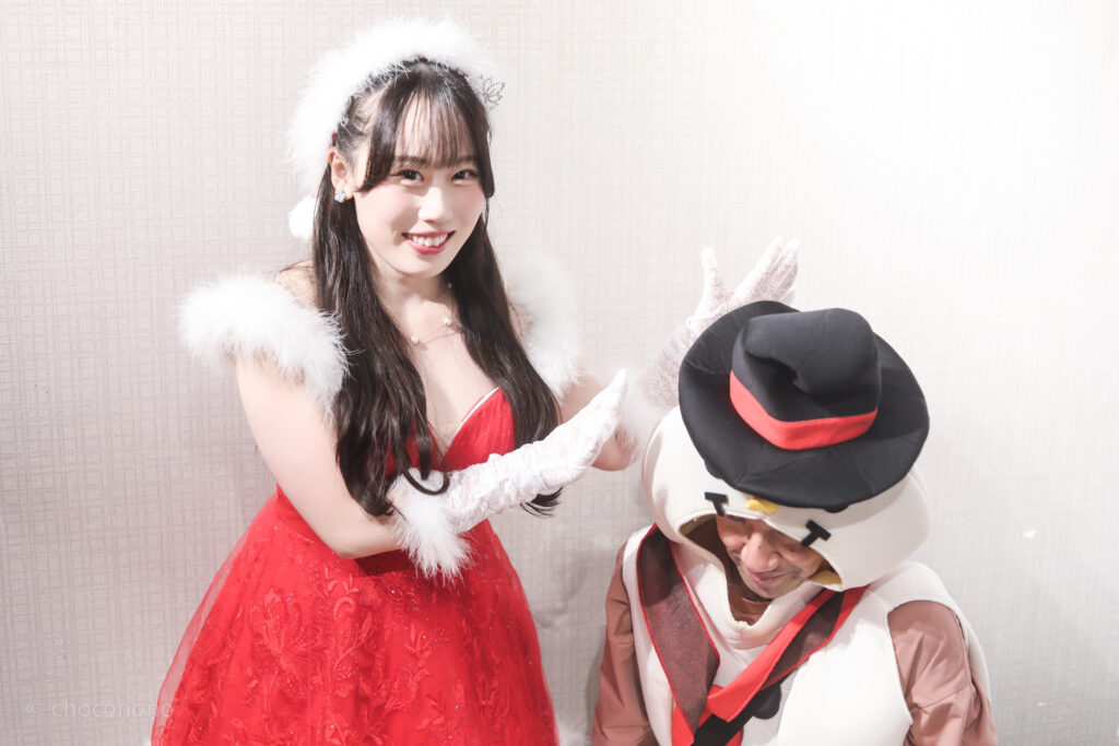 embellir(あんべりー)の甘夏あも(あまなつあも)ちゃんと特典会で撮ってもらったツーショット写真。
あもちゃんは真っ赤なドレス姿にサンタ帽。「大人の雰囲気」だった。
雪だるまの着ぐるみをしていた俺を「作る」ジェスチャー。
2025年　12月25日　冠Fes! Xmas SP!にて撮影