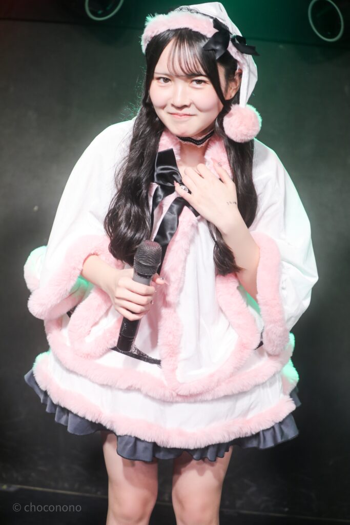 embellir(あんべりー)の遥乃ももか(はるのももか)ちゃんをライブ中に撮影した写真。
ピンクのもこもこのサンタ衣装を着ている。
ちょっと照れているような満更でもないような、絶妙な表情で微笑んでいる。
2025年12月25日GOTANDA G3　冠Fes Xmas SP!にて撮影。