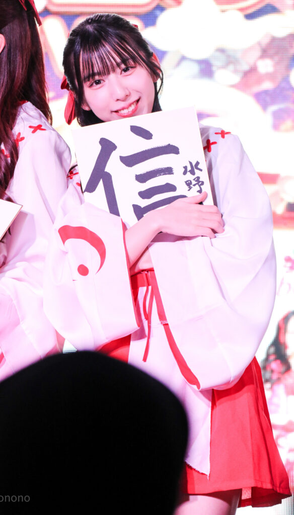 embellir(あんべりー)の水野ひなた(みずのひなた)ちゃんをライブ中に撮影した写真。巫女服を着ている。新春の書き初め企画で書いた信じるの「信」という字を書いた色紙を抱えて、にこにこ笑いながらこちらを見ている。
2026年1月4日SHIBUYA ONE5　新春ONE5LIVEにて撮影。
