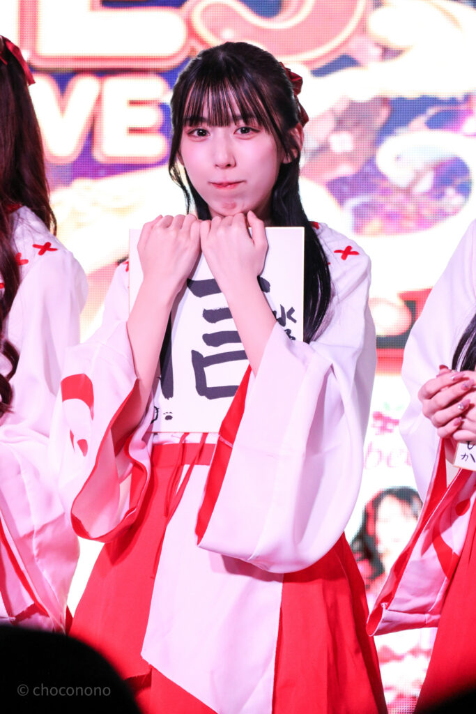 embellir(あんべりー)の水野ひなた(みずのひなた)ちゃんをライブ中に撮影した写真。巫女服を着ている。新春の書き初め企画で書いた信じるの「信」という字を書いた色紙を抱えて、小動物のような可愛らしい表情でこちらを見ている。
2026年1月4日SHIBUYA ONE5　新春ONE5LIVEにて撮影。