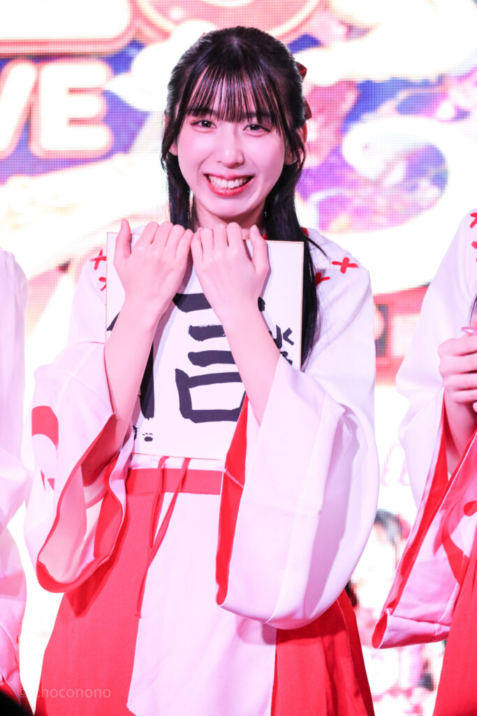 embellir(あんべりー)の水野ひなた(みずのひなた)ちゃんをライブ中に撮影した写真。巫女服を着ている。新春の書き初め企画で書いた信じるの「信」という字を書いた色紙を抱えて、にこにこしながらこちらを見ている。 2026年1月4日SHIBUYA ONE5　新春ONE5LIVEにて撮影。