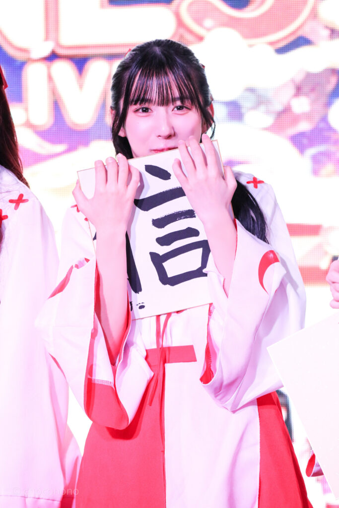 embellir(あんべりー)の水野ひなた(みずのひなた)ちゃんをライブ中に撮影した写真。巫女服を着ている。新春の書き初め企画で書いた信じるの「信」という字を書いた色紙を抱えて、いたずらっぽく笑いながらこちらを見ている。
2026年1月4日SHIBUYA ONE5　新春ONE5LIVEにて撮影。