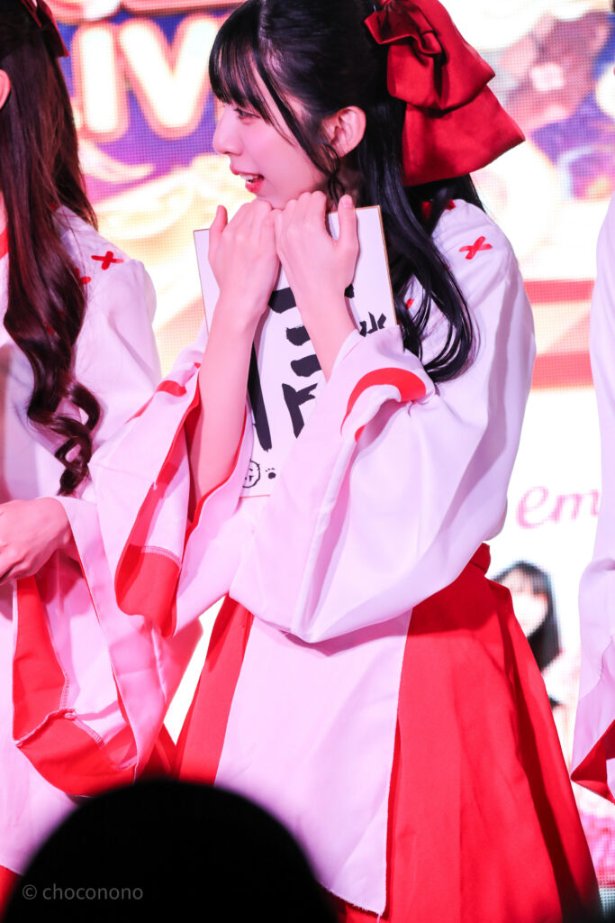 embellir(あんべりー)の水野ひなた(みずのひなた)ちゃんをライブ中に撮影した写真。巫女服を着ている。新春の書き初め企画で書いた信じるの「信」という字を書いた色紙を抱えてステージ反対側のやり取りを見ている。 2026年1月4日SHIBUYA ONE5　新春ONE5LIVEにて撮影。